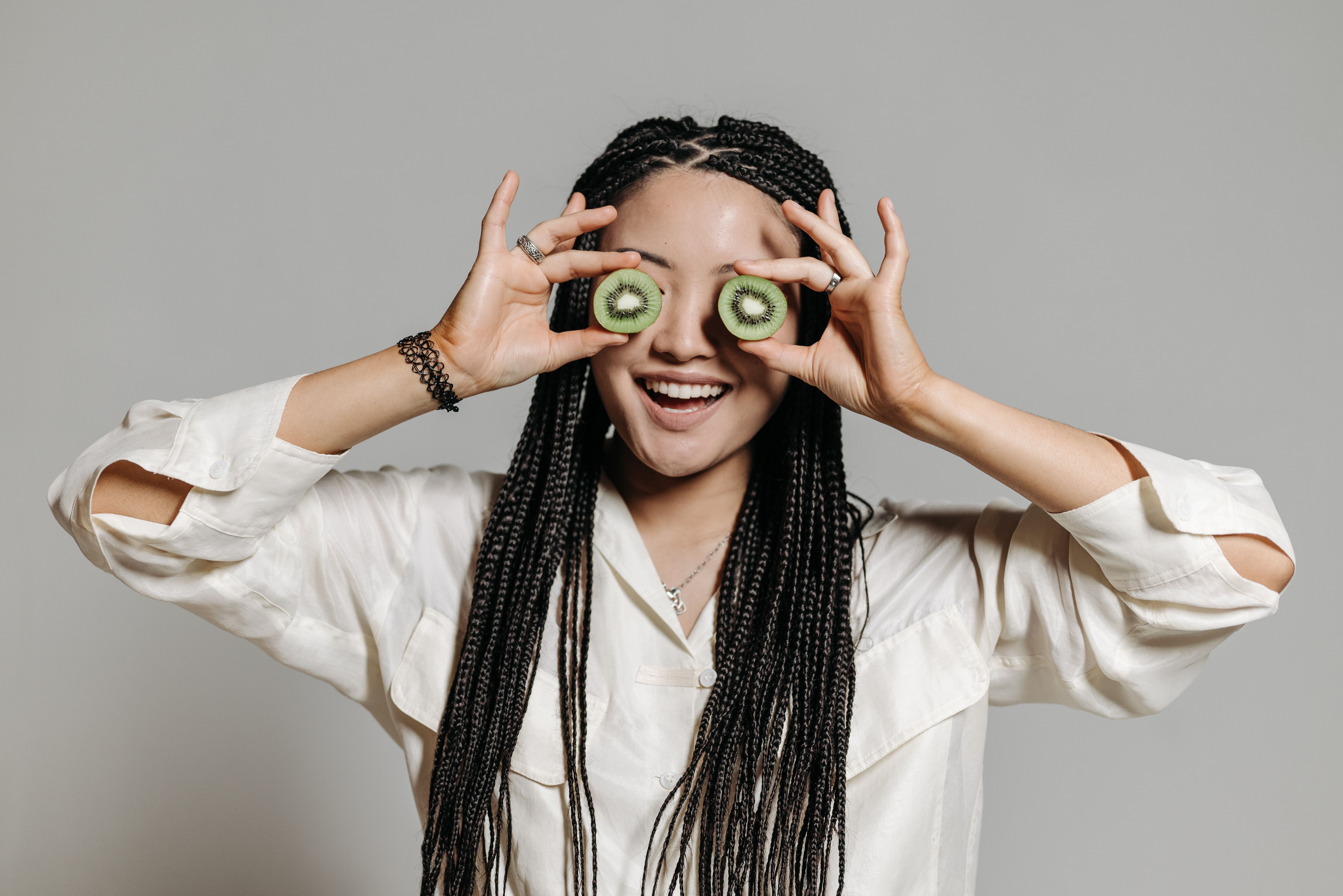 Woman with braided hair playfully holding kiwi slices over her eyes, smiling in a casual setting.