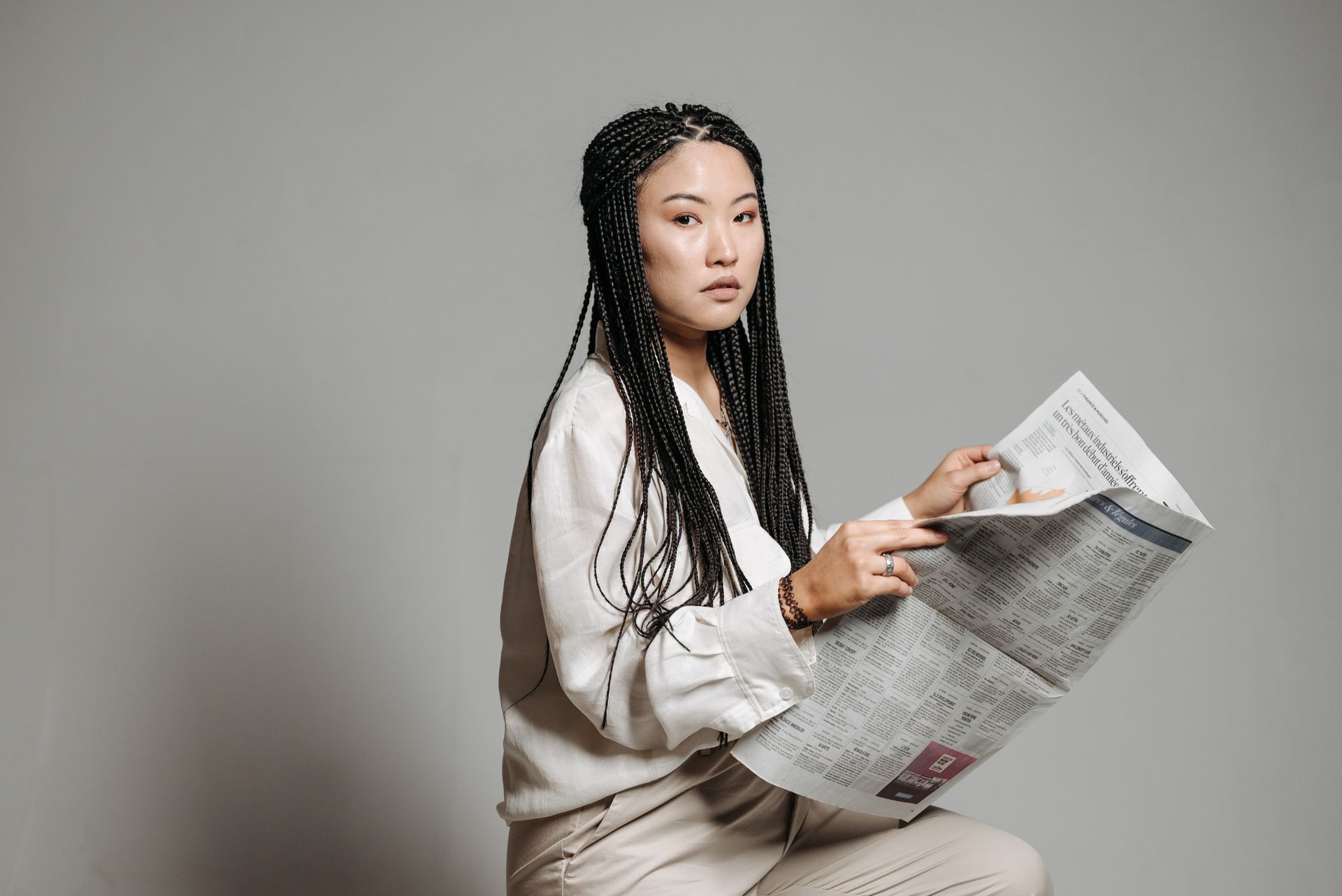 Asian woman with braided hair reads a newspaper, wearing a button-down shirt in a studio setting.