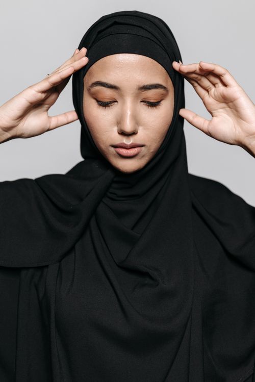 Free Portrait of a young woman wearing a black hijab, hands gracefully on her head, eyes closed in a calm pose. Stock Photo