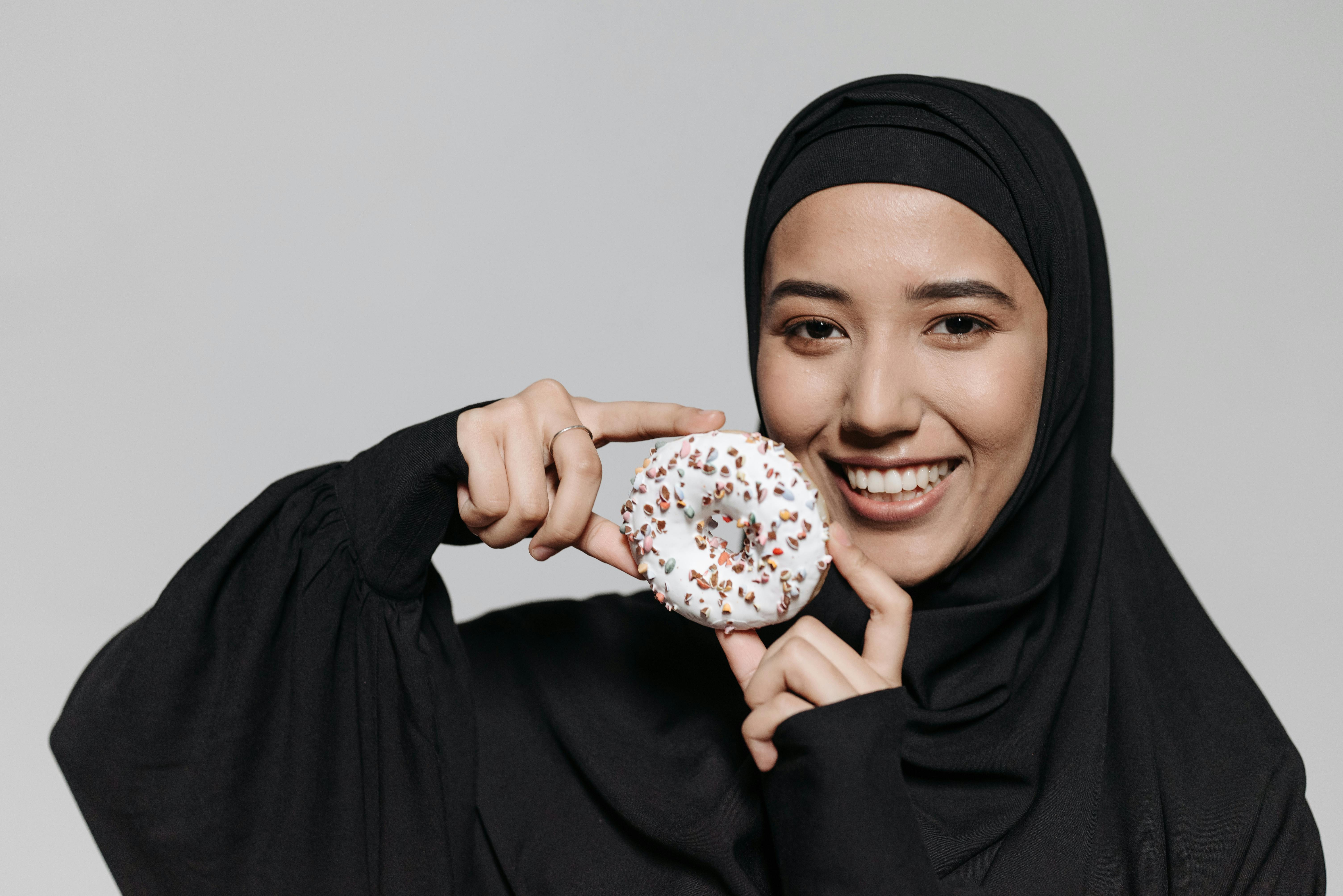 black kimono abaya - Smiling young woman in a hijab holding a donut and posing joyfully for a portrait shot.