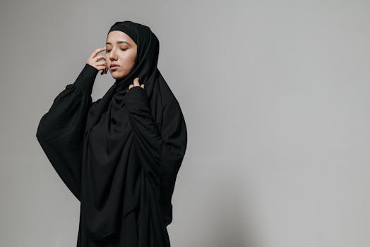 A serene portrait of a young woman wearing a black hijab and abaya against a neutral background.