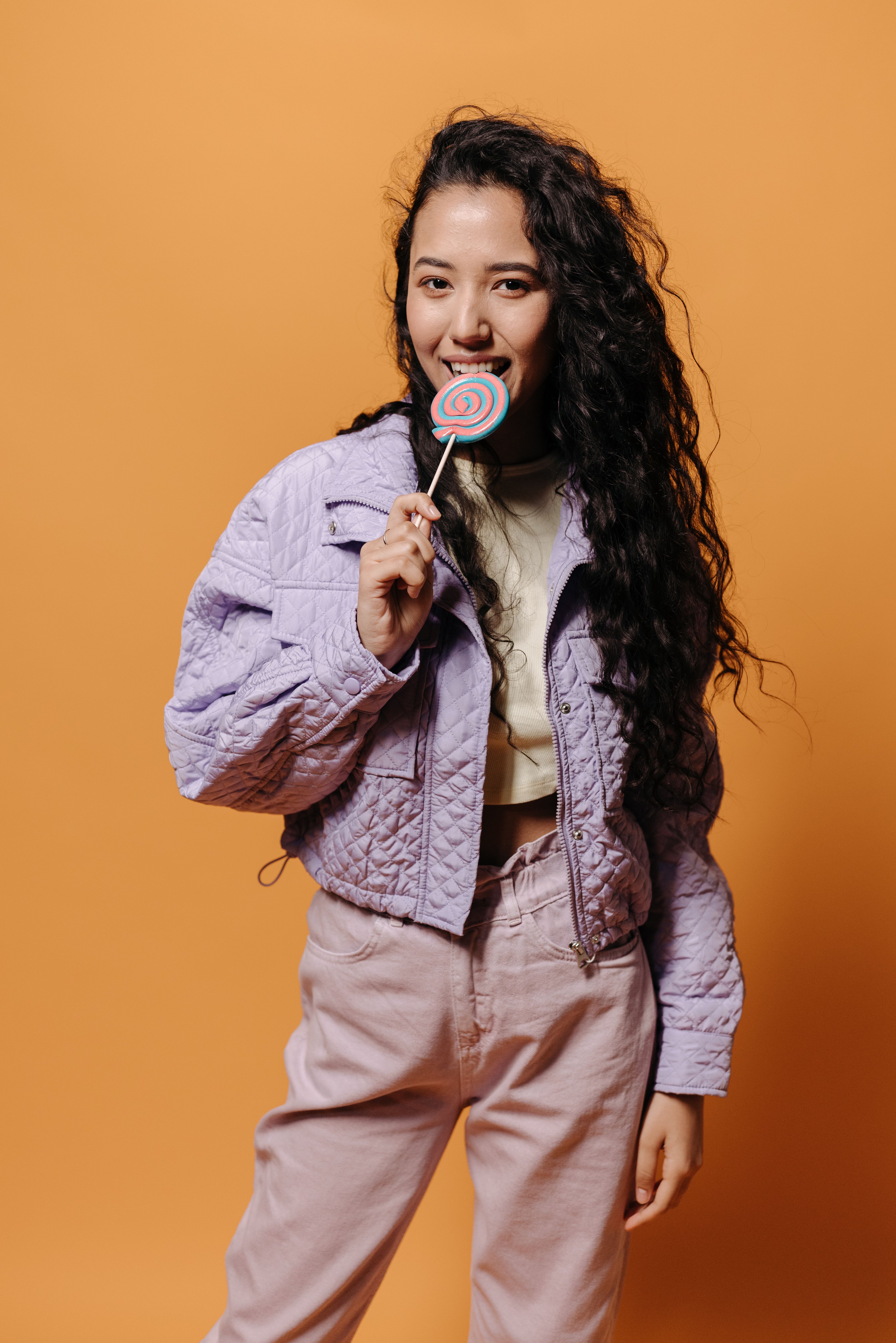 Stylish young woman in a purple jacket enjoying a lollipop against a peach background.
