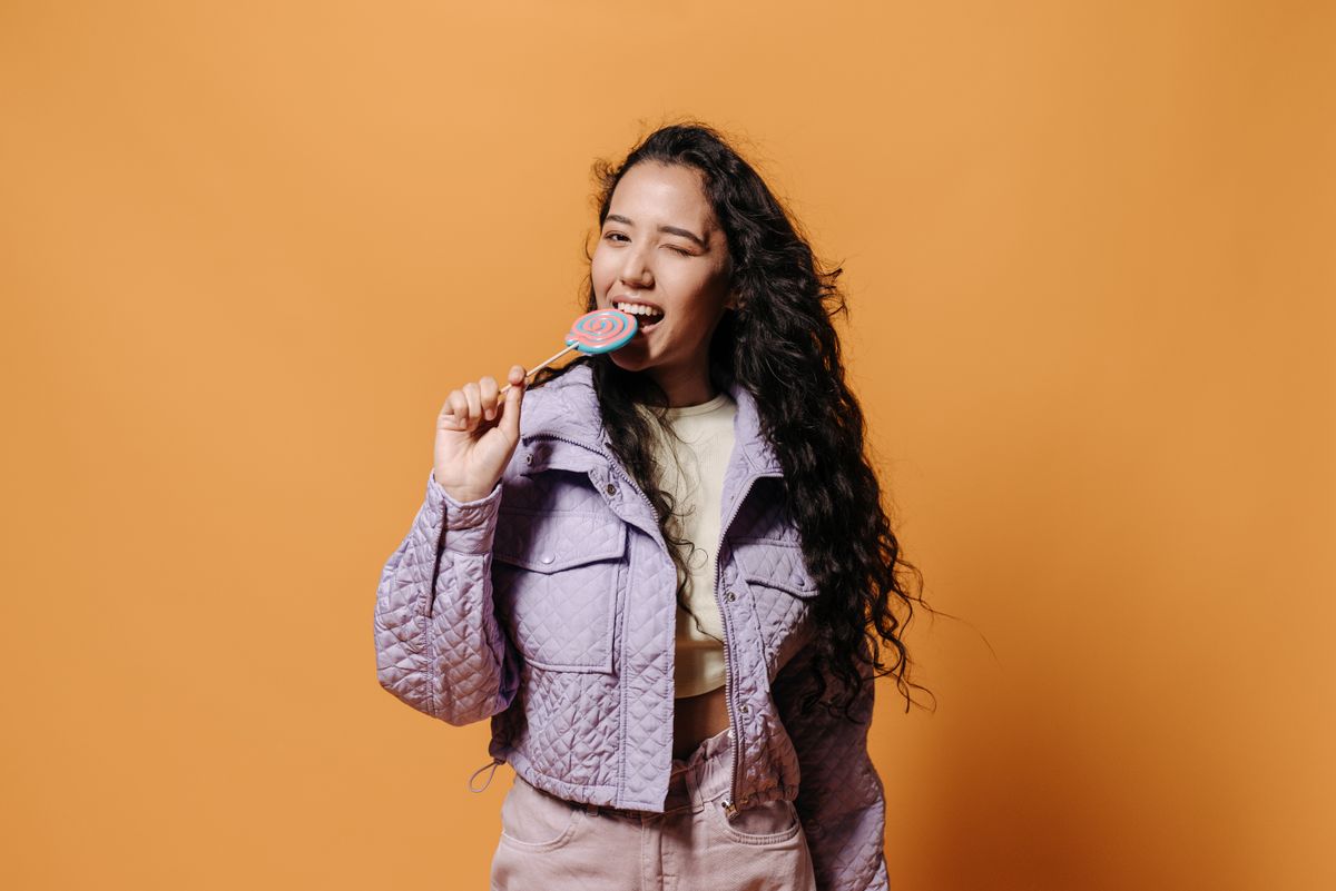 Cheerful woman in purple jacket biting a colorful lollipop and winking on an orange backdrop.