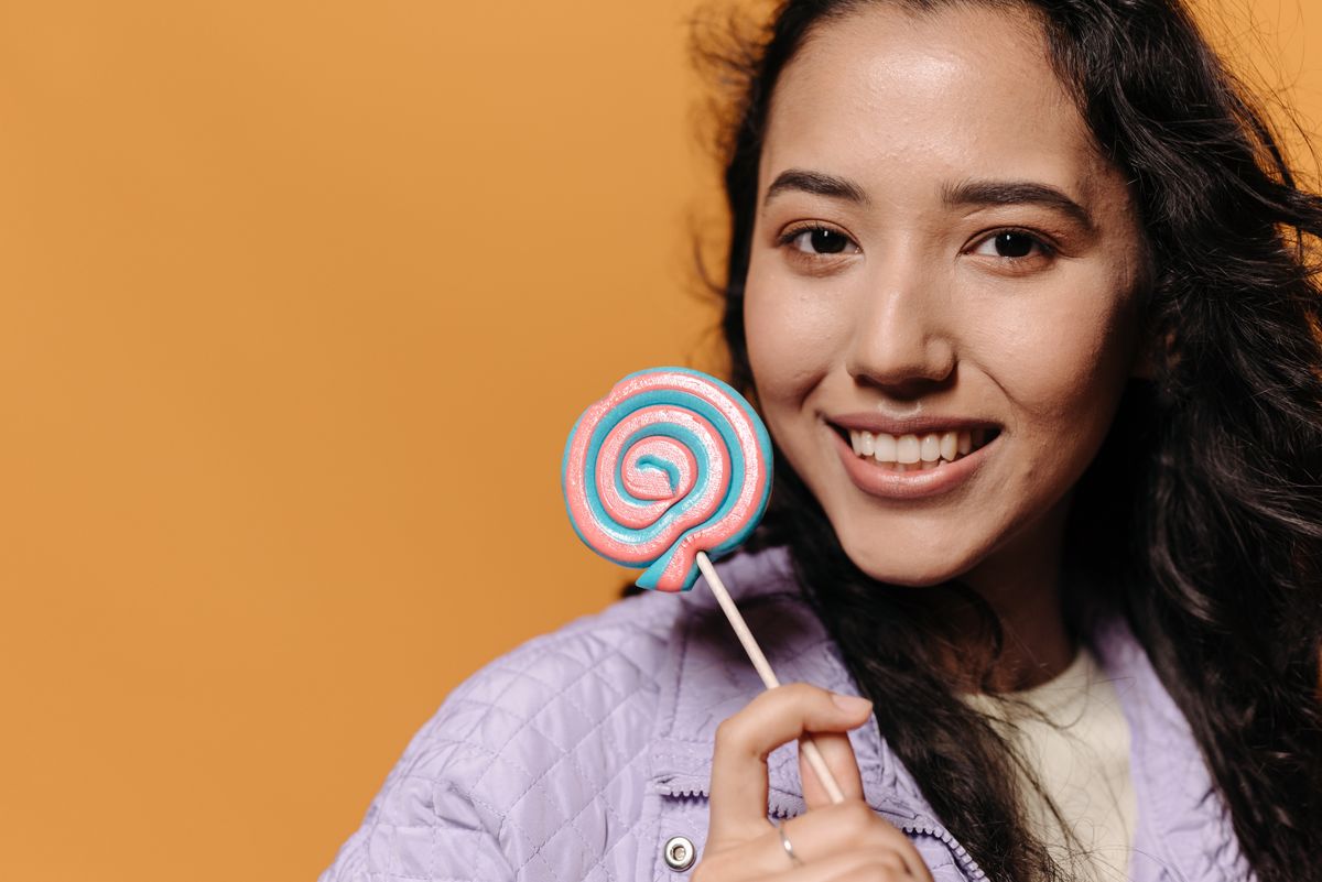 Portrait of a smiling young woman holding a vibrant spiral lollipop on a peach background.