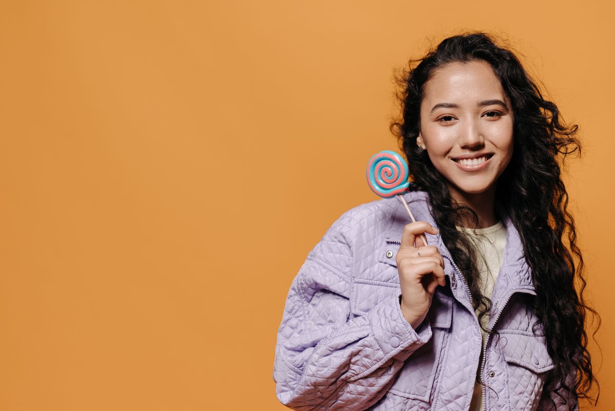 Cheerful woman with curly hair holds a colorful lollipop against an amber background.