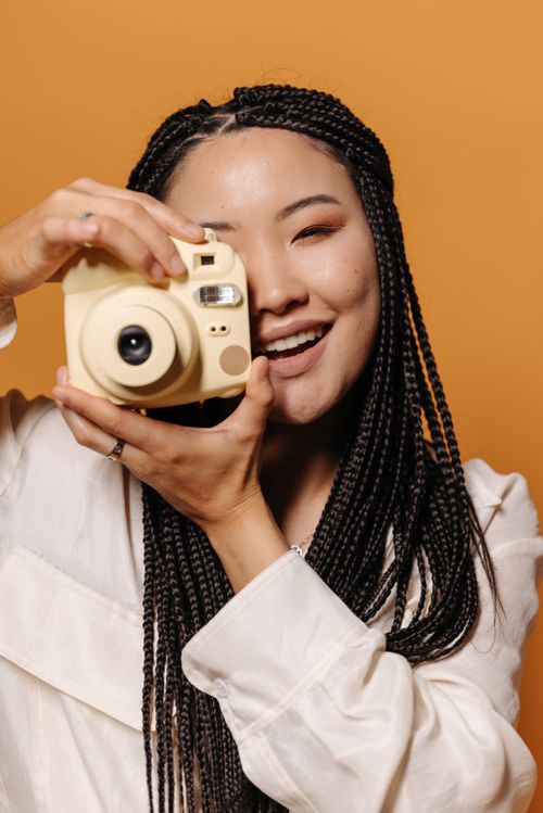 Free Asian woman smiling while taking a photo with an instant camera against an orange backdrop. Stock Photo