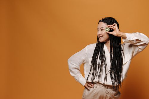 Free Stylish Asian woman with braided hair posing happily in a studio with an orange background, holding a kiwi fruit. Stock Photo