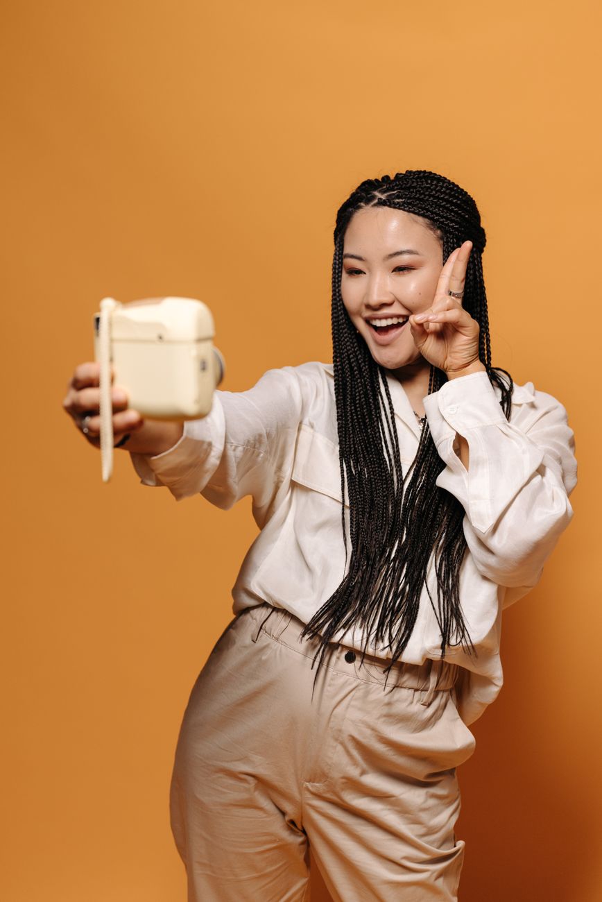 Asian woman with braided hair taking a selfie, posing with a peace sign on an orange background.