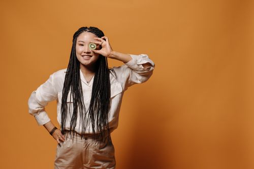 Free Portrait of a young woman holding a kiwi slice against an orange background, smiling joyfully. Stock Photo