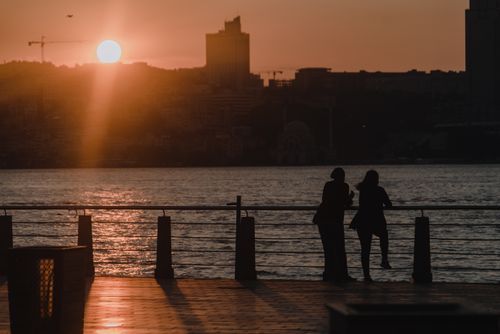 Free Two people silhouetted against a sunset by the river, with urban skyline in the background. Stock Photo