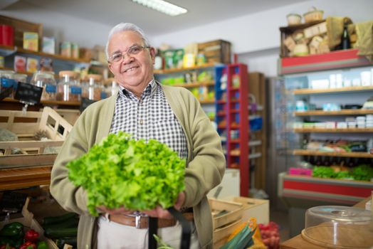 Elderly man with gray hair holding fresh lettuce smiling in a grocery store.