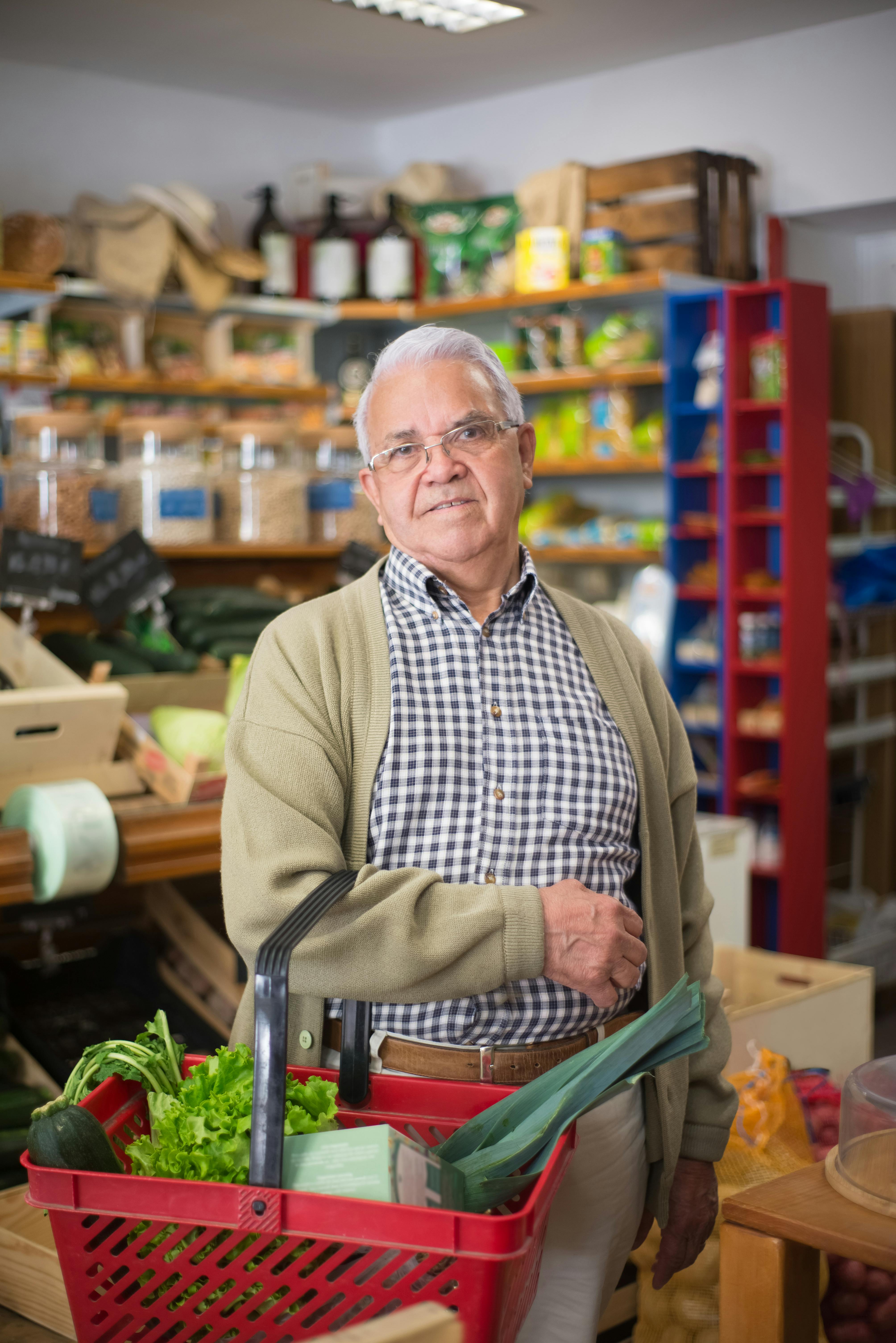 Elderly man shopping in a grocery store with a basket of fresh vegetables.