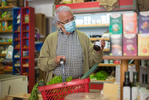 Elderly man with mask shopping for groceries indoors, carrying a basket.