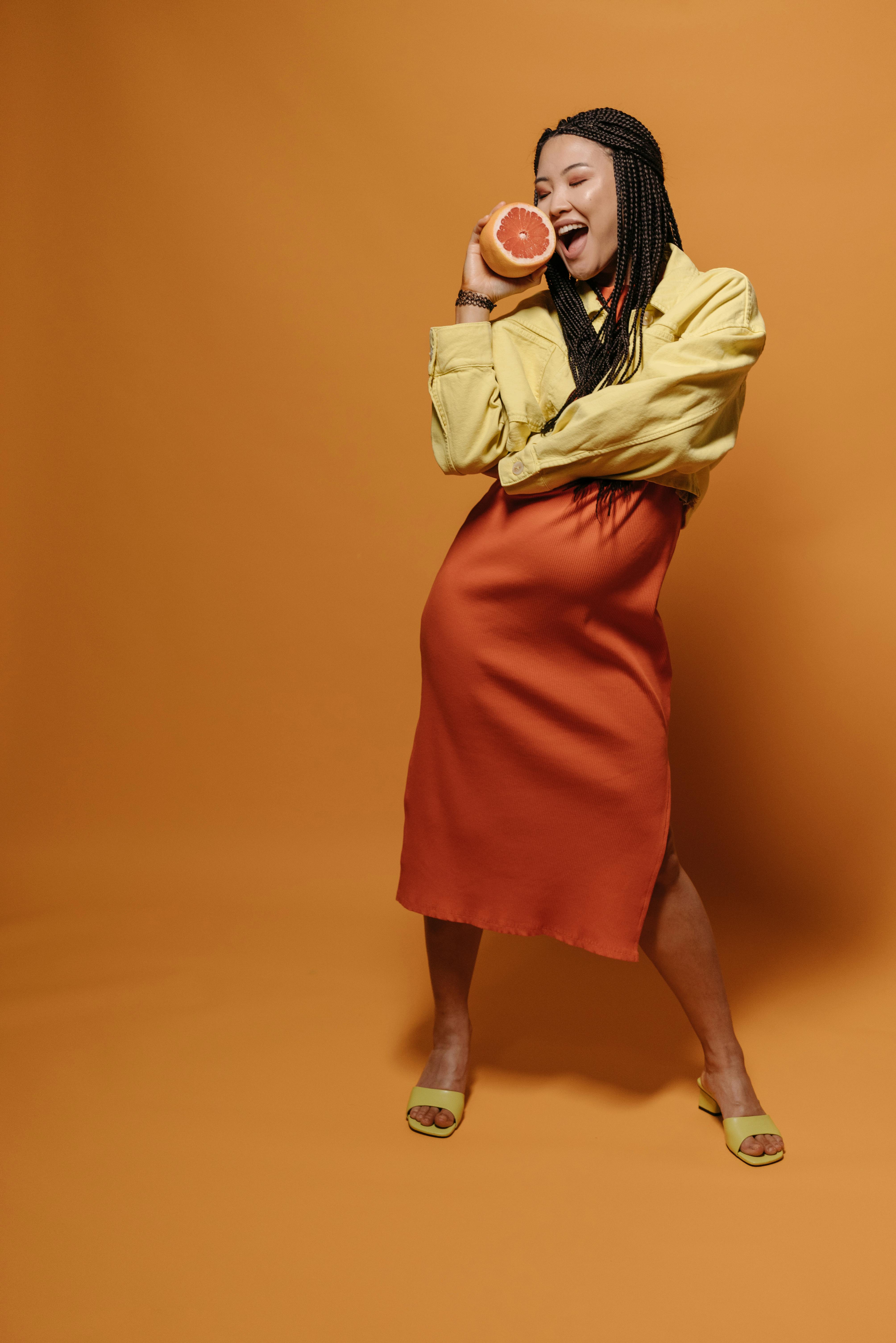 Energetic woman with braided hair holds a grapefruit, posing in a colorful studio setting.