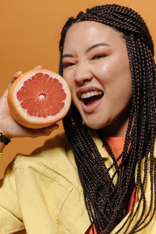 Free Smiling woman with braided hair holding a juicy pomelo slice against an orange background. Stock Photo