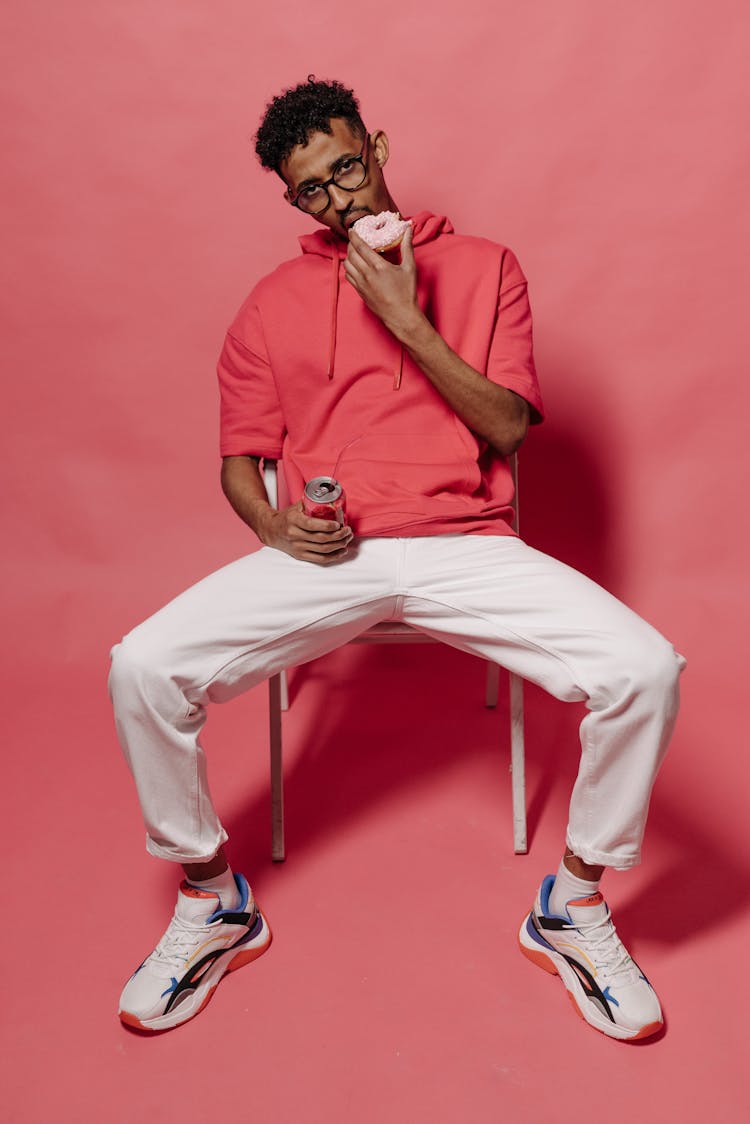 Man In Pink Hoodie And White Pants Sitting On A Chair While Eating Snacks