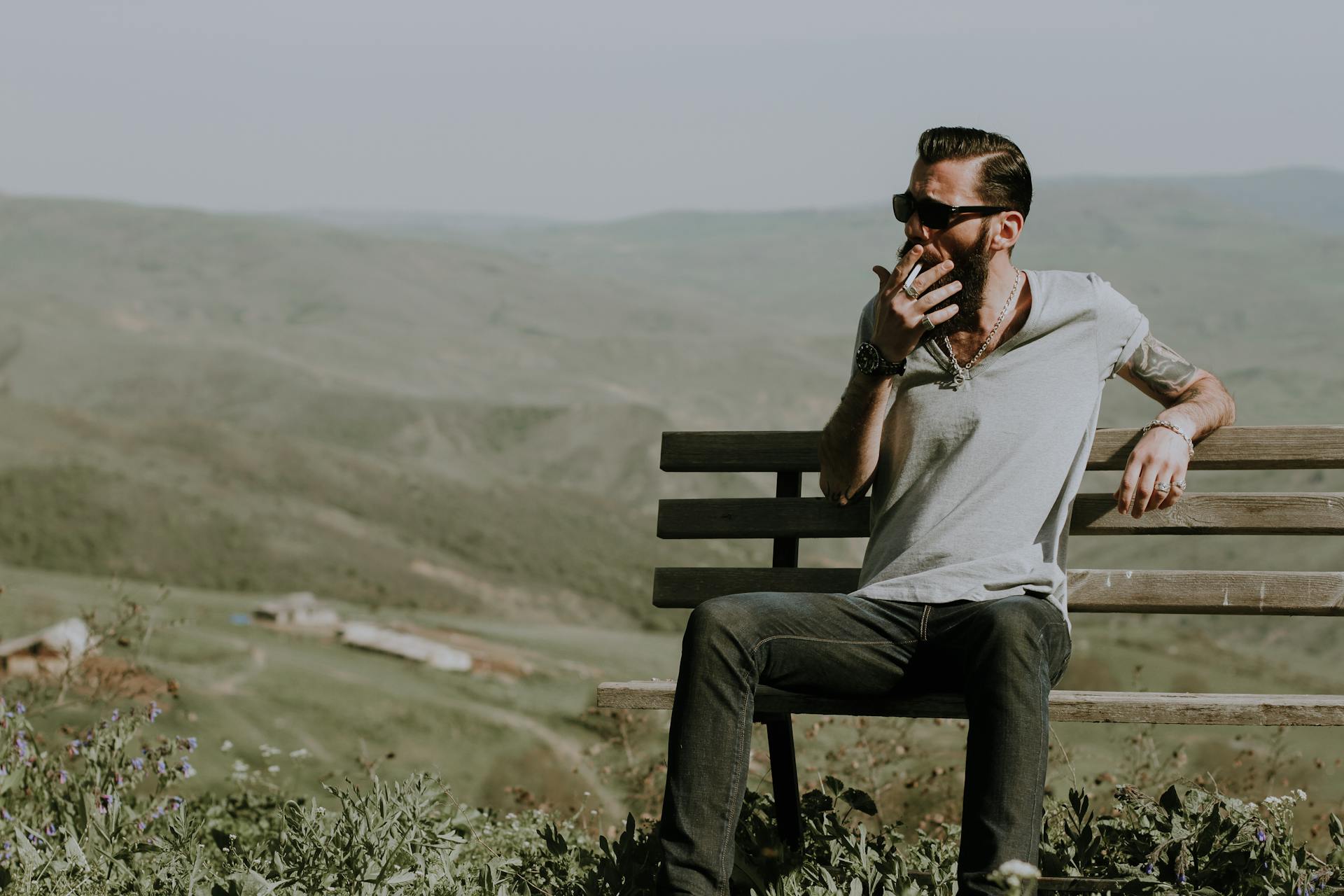 Bearded man with sunglasses and tattoos enjoys a cigarette on a bench amidst scenic Azerbaijan mountains.