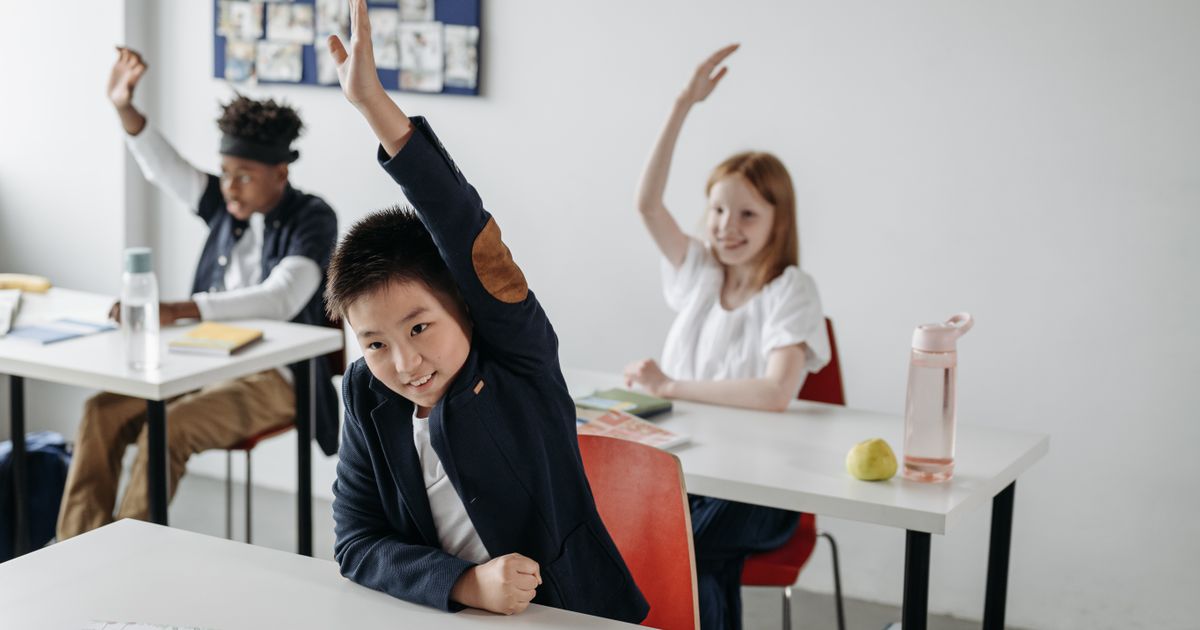 Elementary students seated at classroom tables raising their hands during a lesson