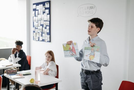 A young student in a classroom presenting colorful drawings to classmates during an art lesson.