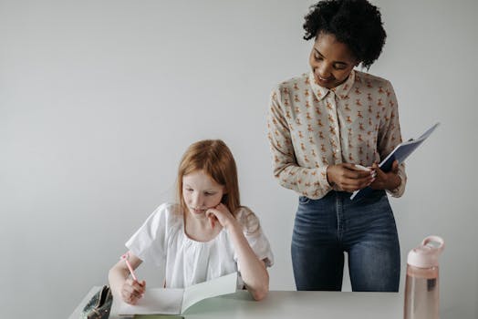 A teacher guides a student with her writing assignment at a classroom desk.