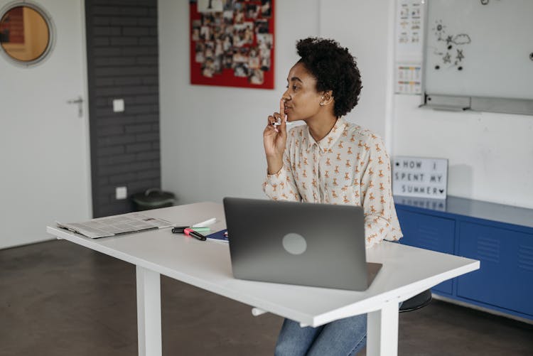 A Woman Sitting At A Table With Laptop Doing A Hush Hand Gesture