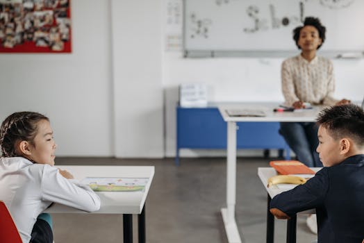 Two students engage in a classroom setting with their teacher in the background.
