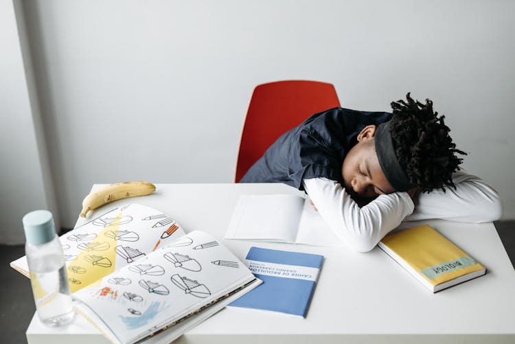 A Kid Sleeping On White Desk With Textbooks