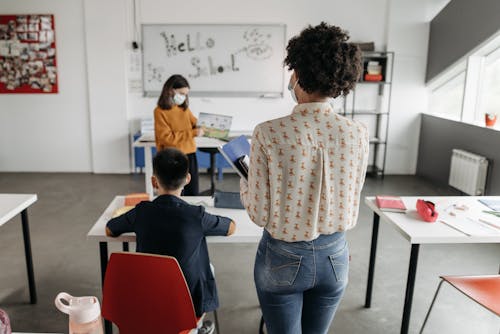 A Person Standing Beside a Kid Sitting at a Desk