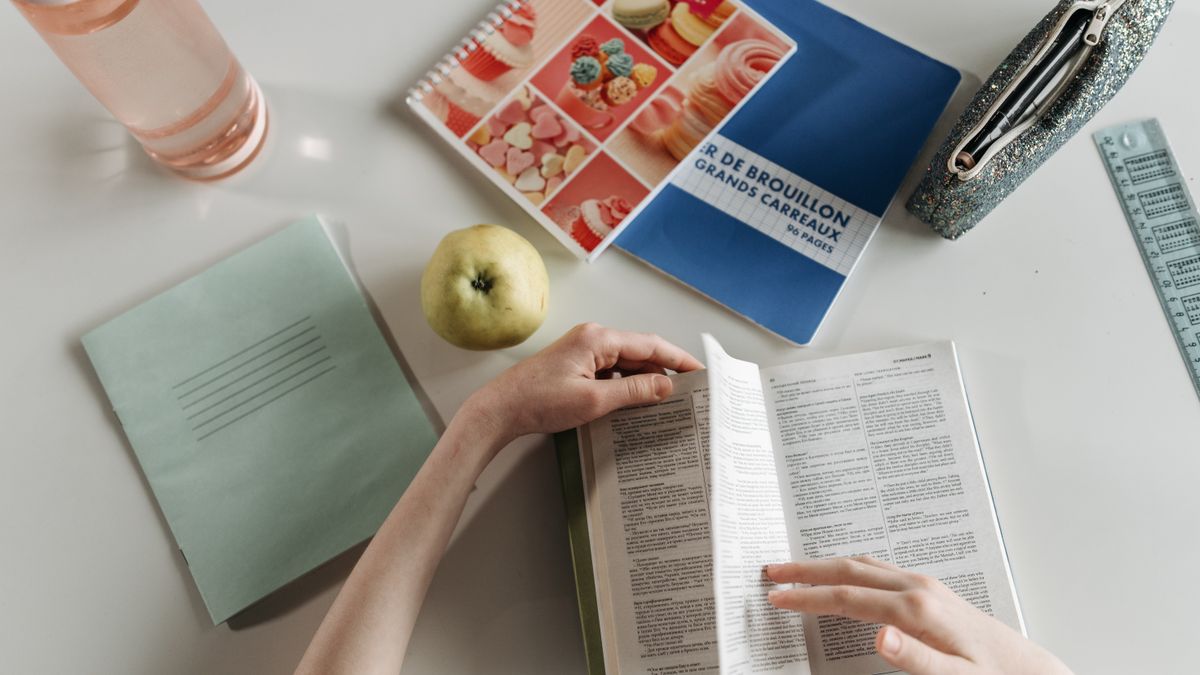 Overhead view of hands turning book pages at a study desk with notebooks, apple, and water bottle.