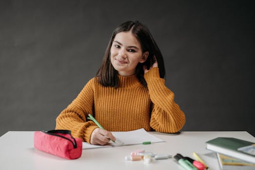 A cheerful student wearing a brown sweater sits at a desk with school supplies in a classroom.