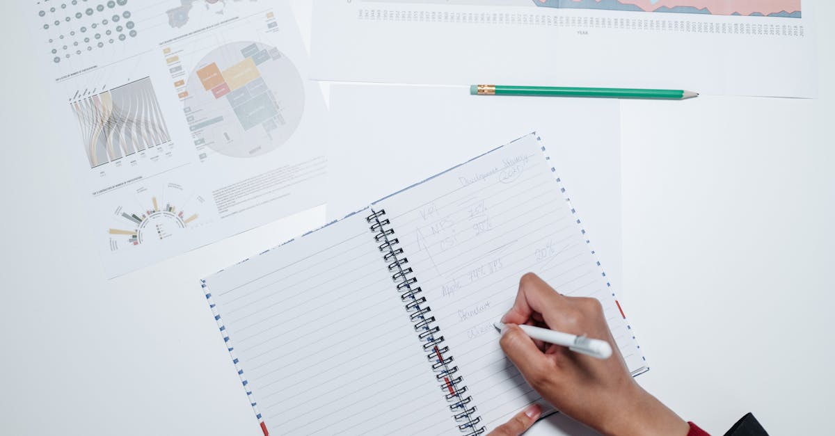 Hands writing in a notebook on a desk with charts, indicating office work.