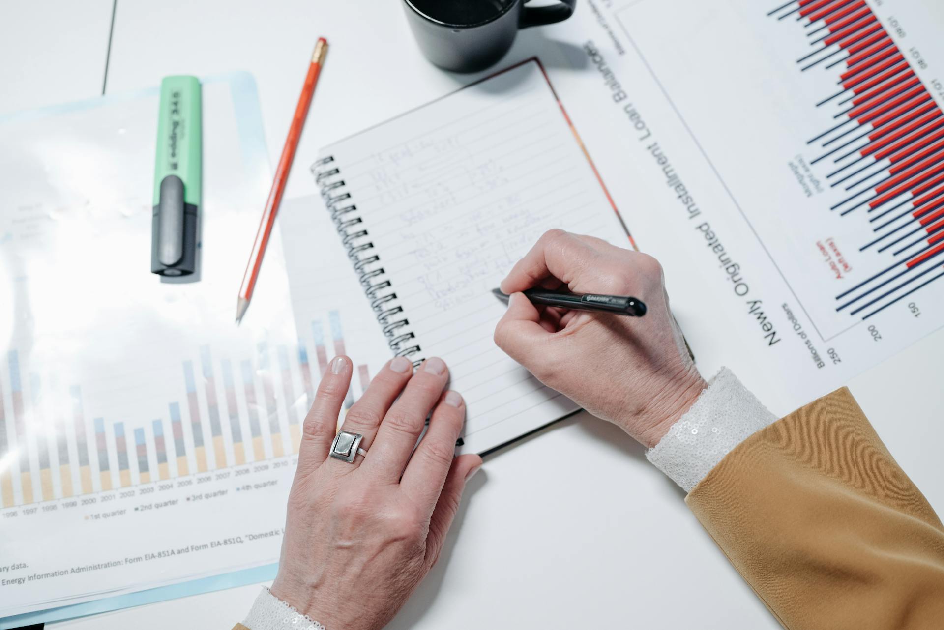 Close-up of hands writing notes in a notebook with charts and graphs on a desk.