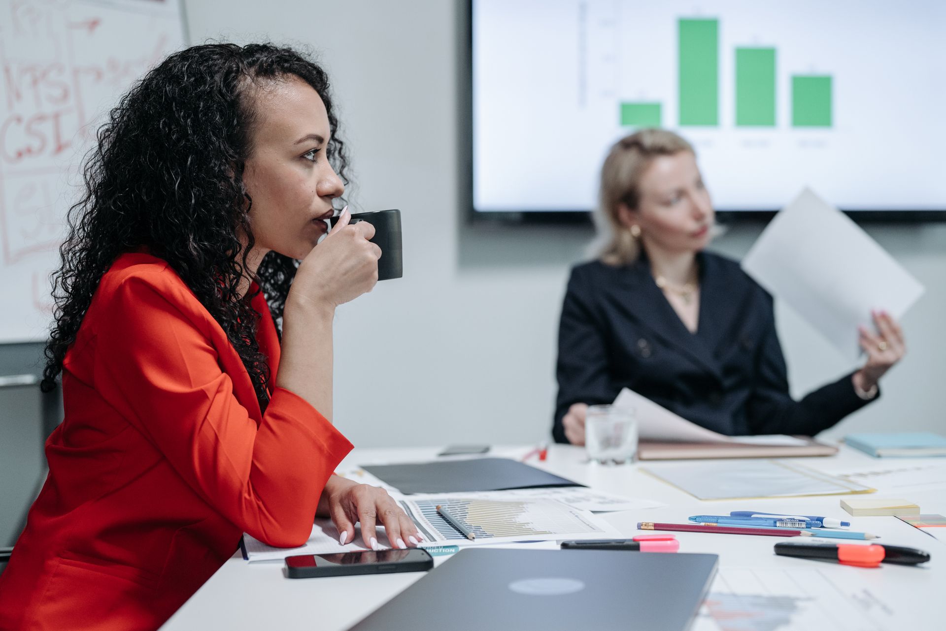 Two women in a business meeting, discussing documents and charts, with focus on one drinking coffee.