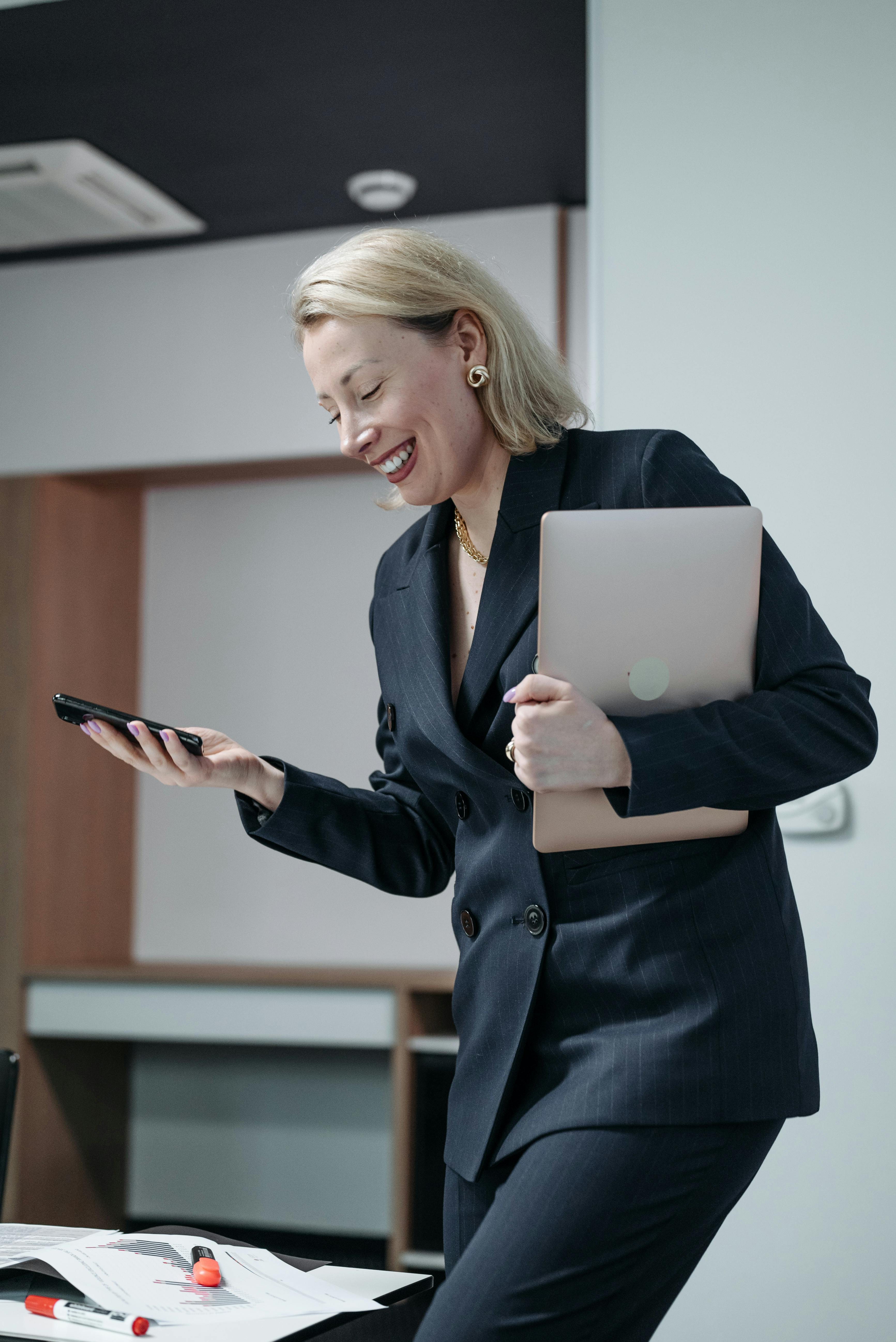 woman in black blazer holding a laptop while using a smartphone