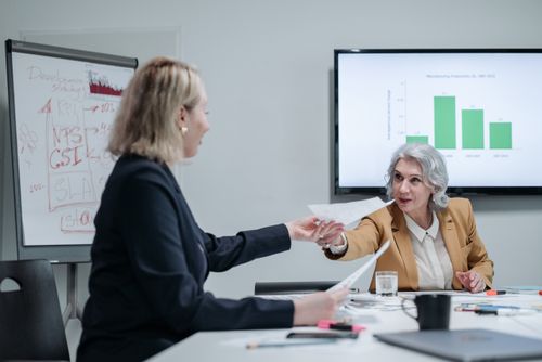 Free Two businesswomen exchanging documents in a modern office setting with presentation screens. Stock Photo