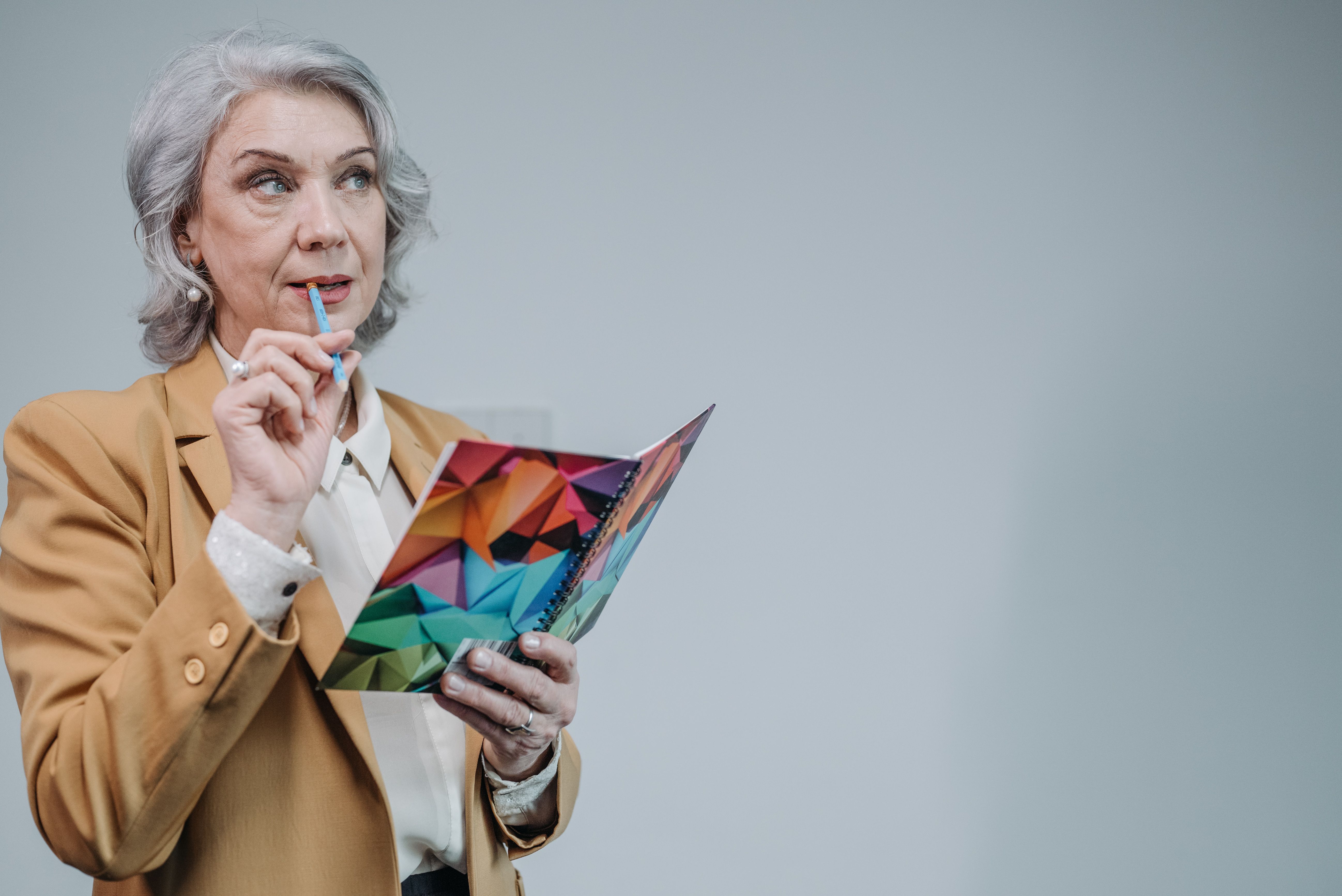 Elderly woman in brown blazer thinking while holding a colorful notebook against a gray background.