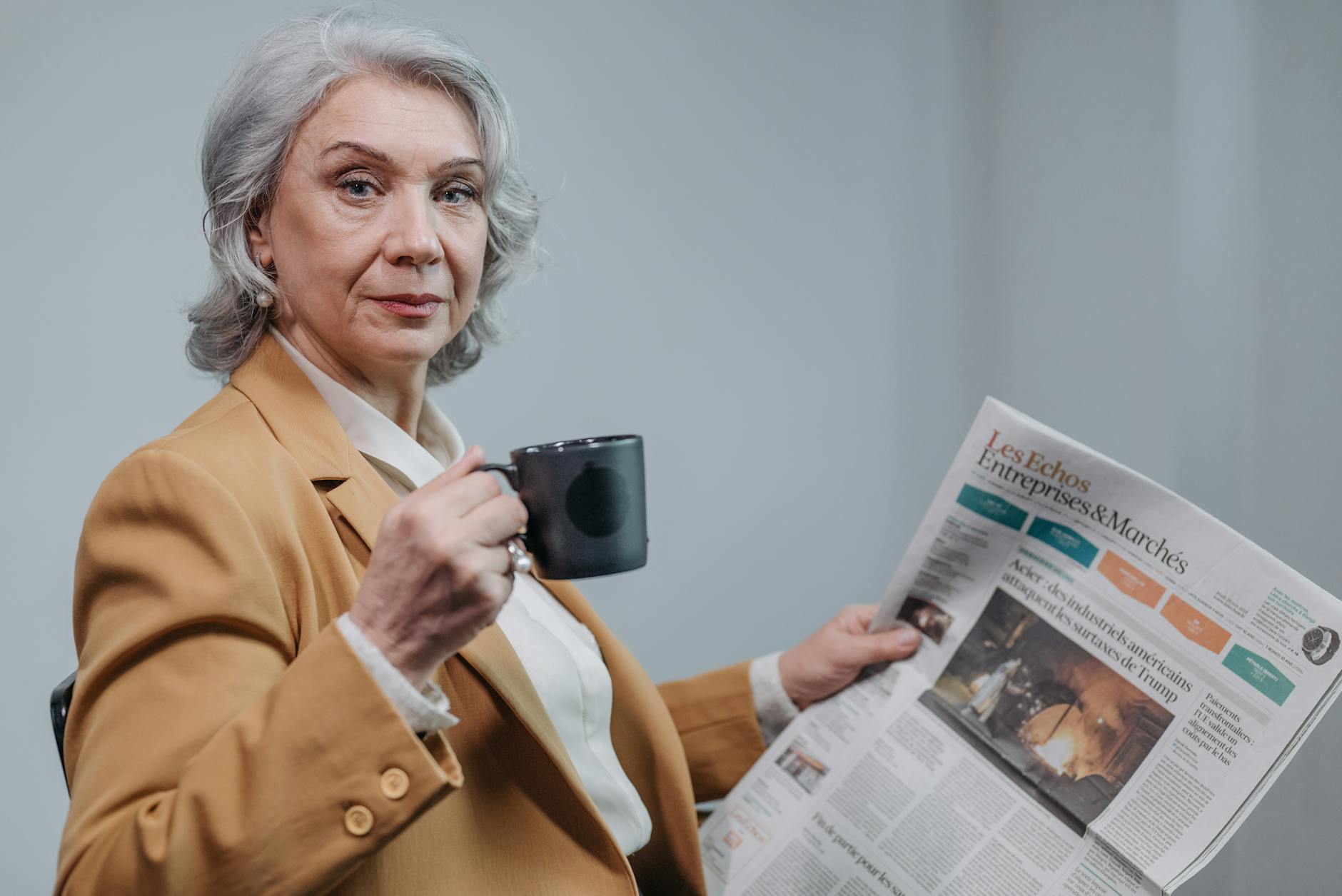 Confident senior businesswoman holding coffee and reading a newspaper indoors.