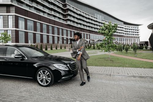 Free Confident man in suit poses beside luxury car with modern building backdrop outdoors. Stock Photo