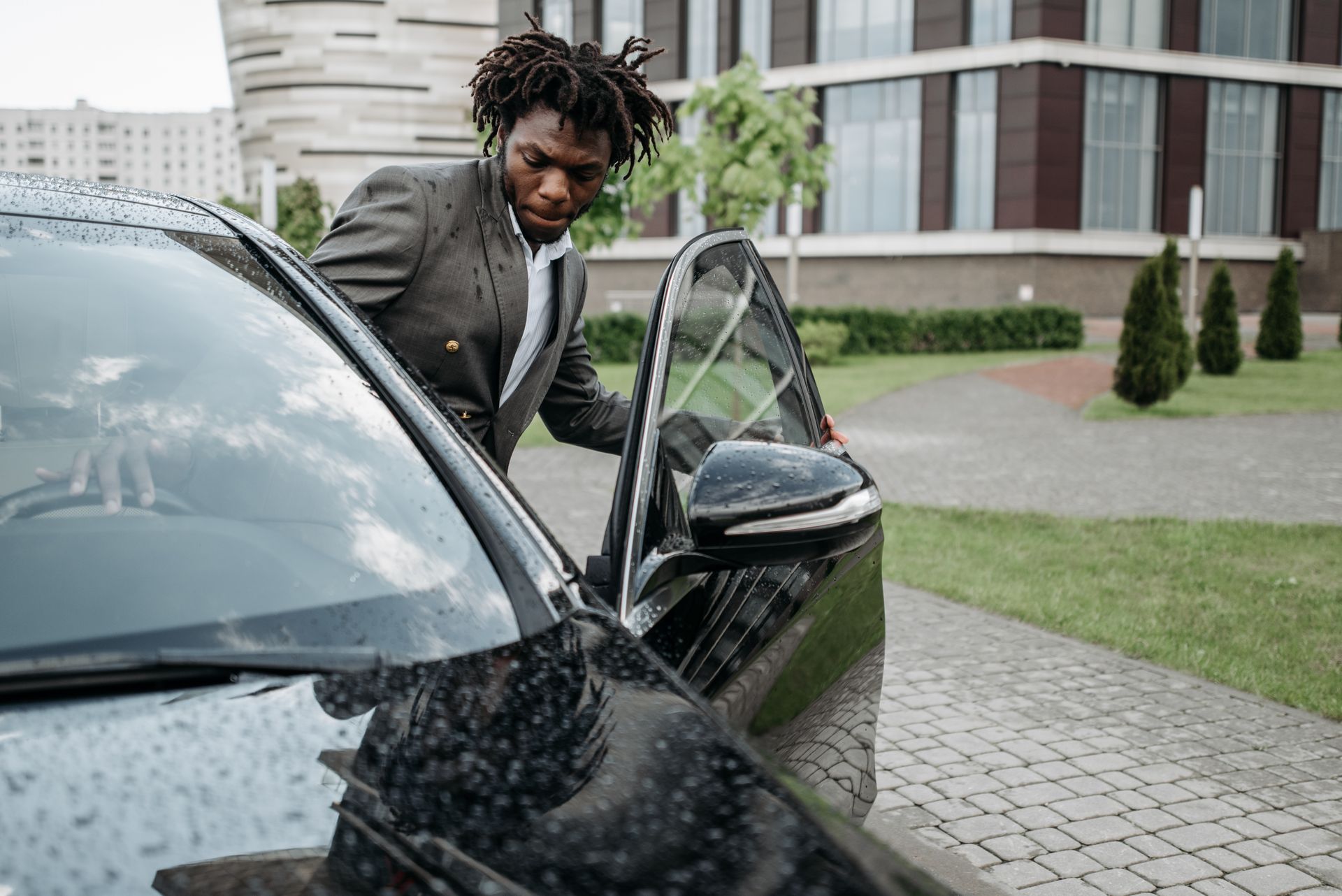 An African American man in a gray suit exits a black car in front of a modern office building on a rainy day.