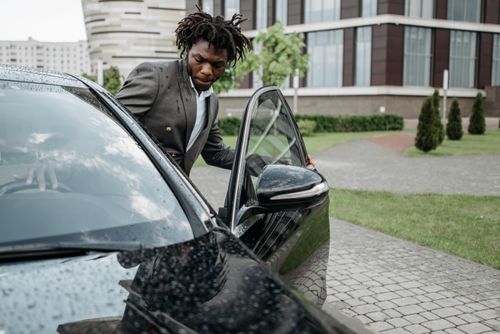 Free An African American man in a gray suit exits a black car in front of a modern office building on a rainy day. Stock Photo
