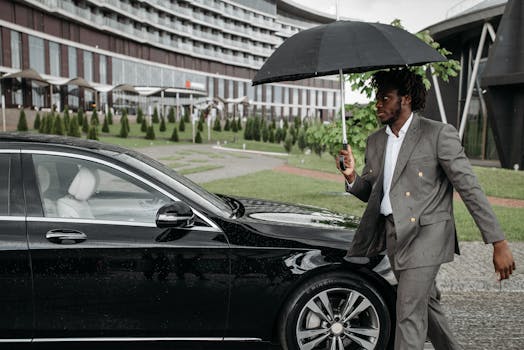 African American man in a suit walking by a car under an umbrella on a rainy day.
