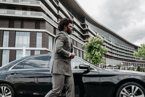 Free Confident businessman in suit walks next to a sleek, black luxury car and modern building. Stock Photo