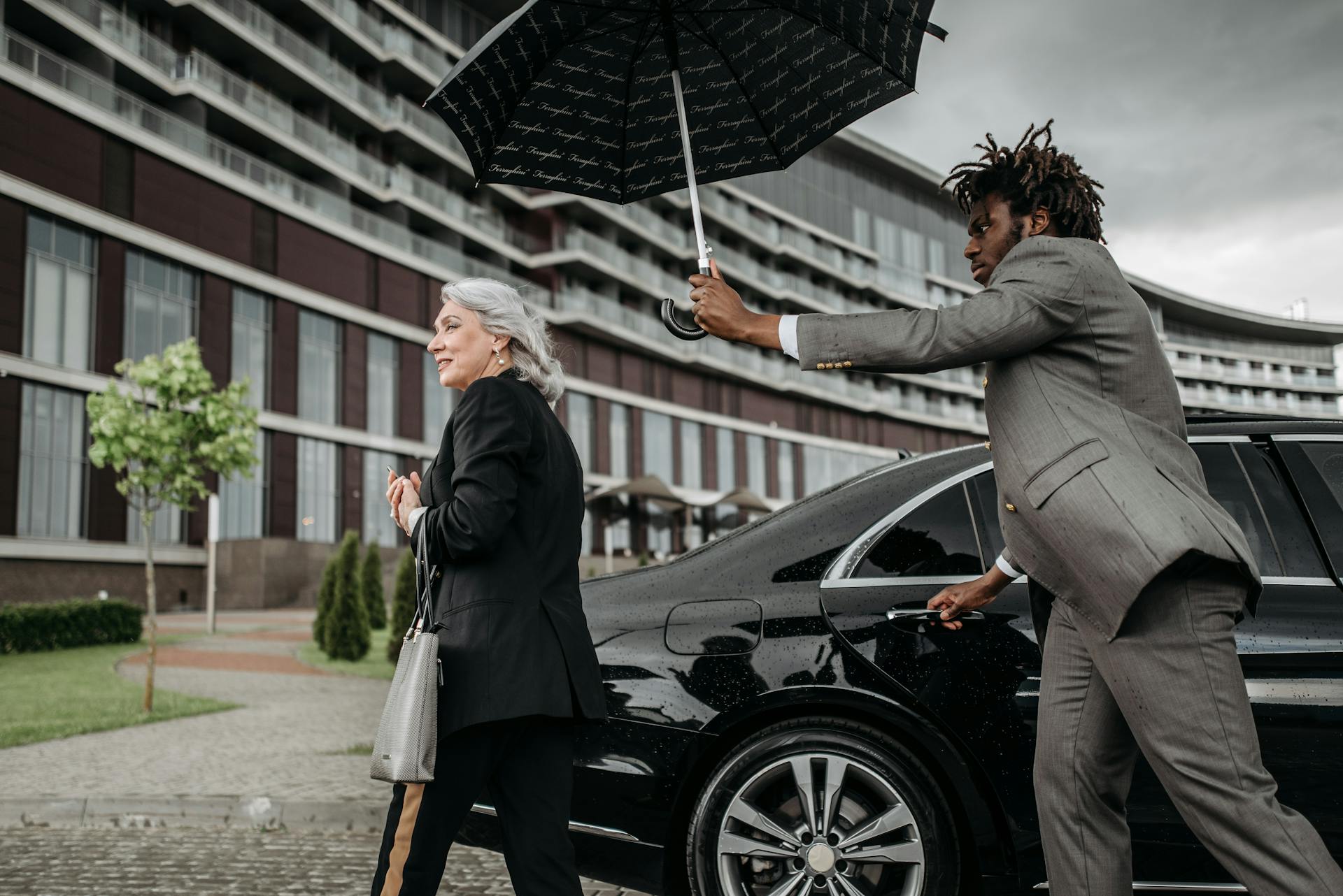 A business professional holds an umbrella for a colleague as they exit a luxury car in front of an office building.