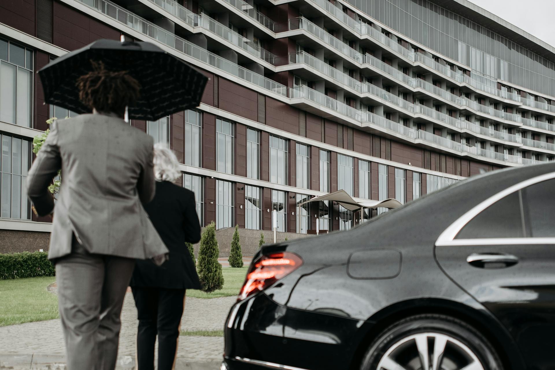 Man in Brown Suit Holding Black Umbrella at the Back of a Senior Woman Walking in Black Blazer