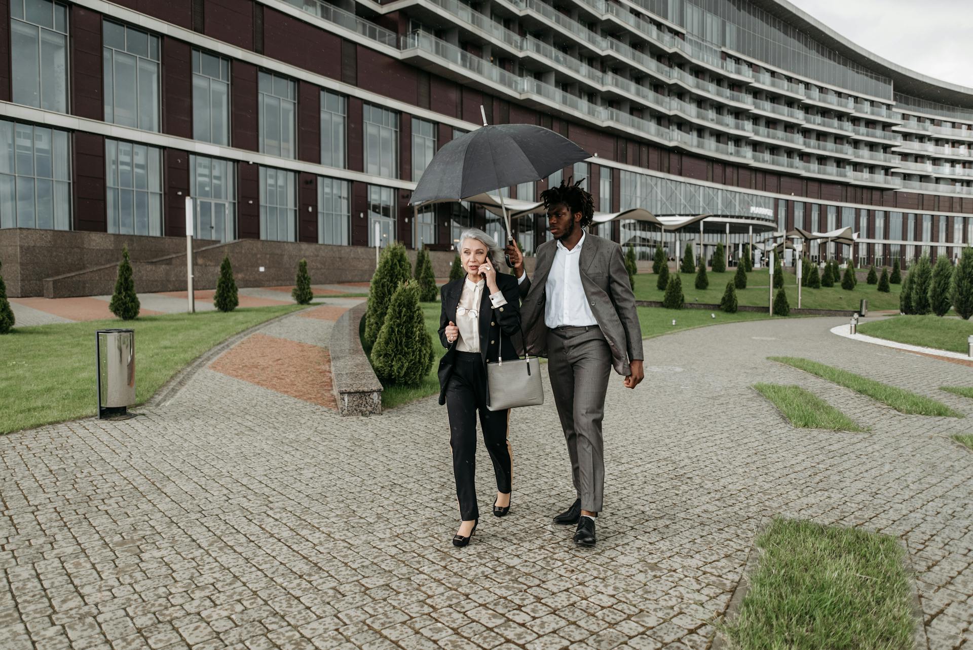 Two business professionals holding an umbrella and walking outside a modern building.