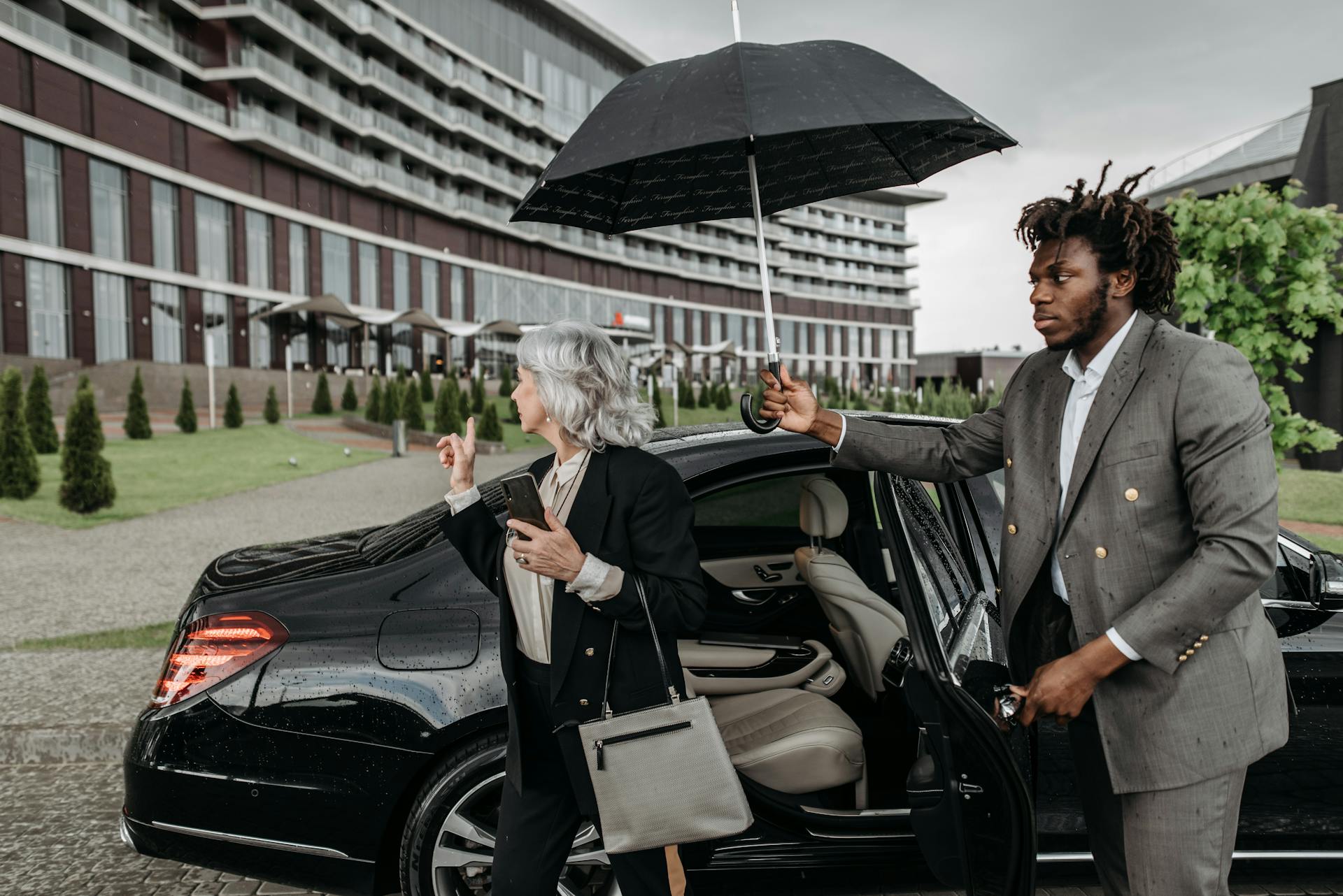 Business professionals exiting a luxury car as an assistant holds an umbrella on a rainy day.