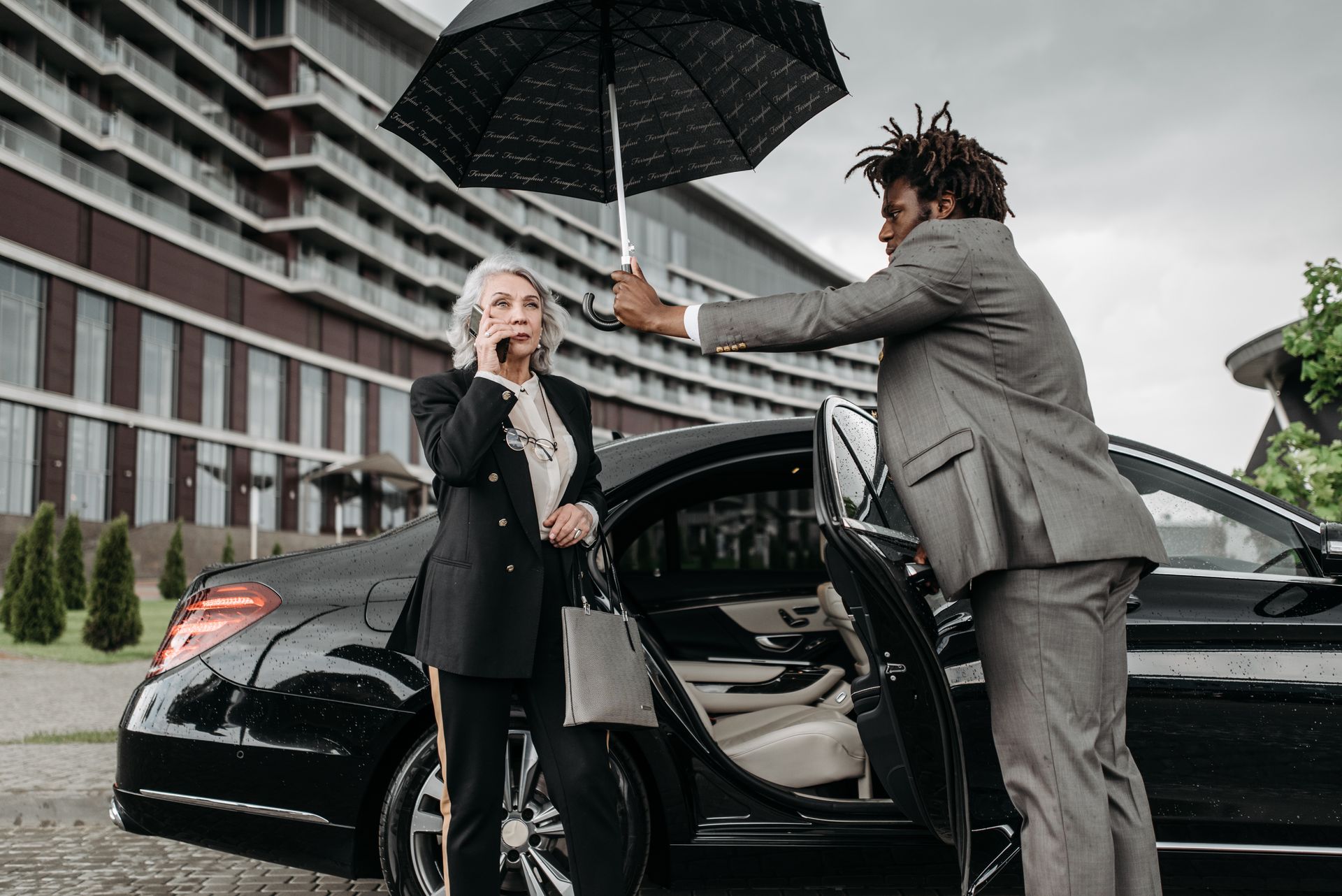 A Woman in Black Blazer Talking on the Phone while Standing Beside Her Assistant Holding an Umbrella