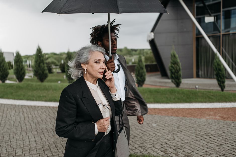 Senior businesswoman talking on phone with umbrella, outdoors alongside a colleague.