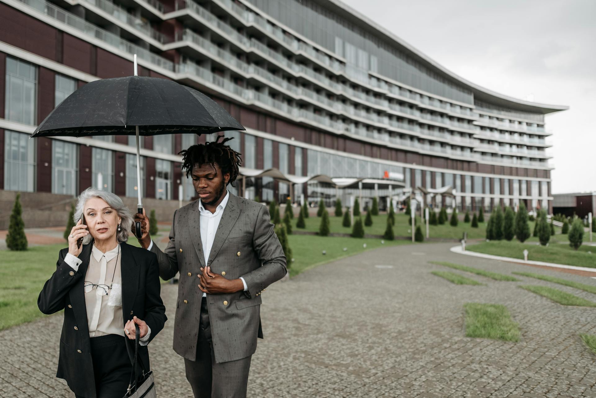 Two business professionals walking outdoors, one holding an umbrella, near a modern building on a cloudy day.