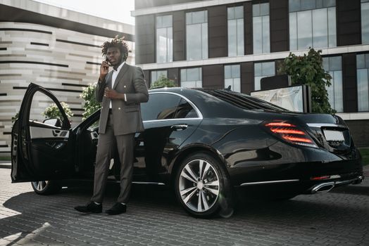 Elegantly dressed man in gray suit making phone call next to sleek black car.